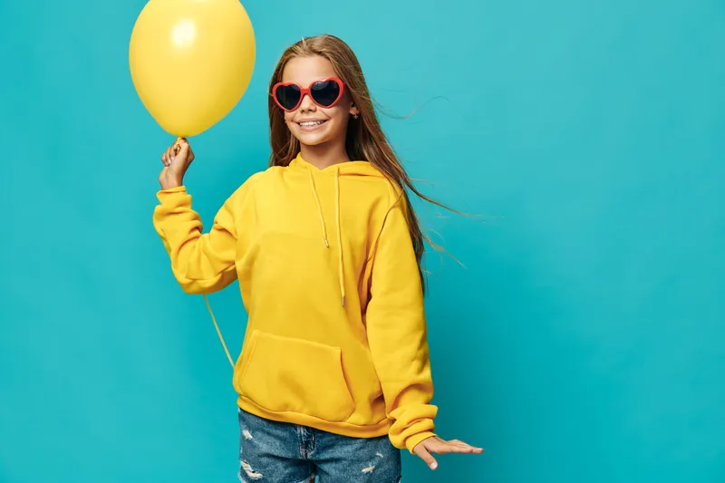 Girl in yellow hoodie holding a balloon with a smile and heart-shaped sunglasses on blue background. Child enjoying party celebration and colorful fashion style outdoors.