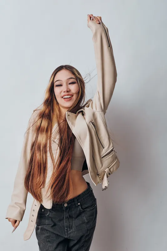 Young woman with long hair smiling and raising hand wearing beige jacket and black jeans posing on white background in studio, casual style and confidence