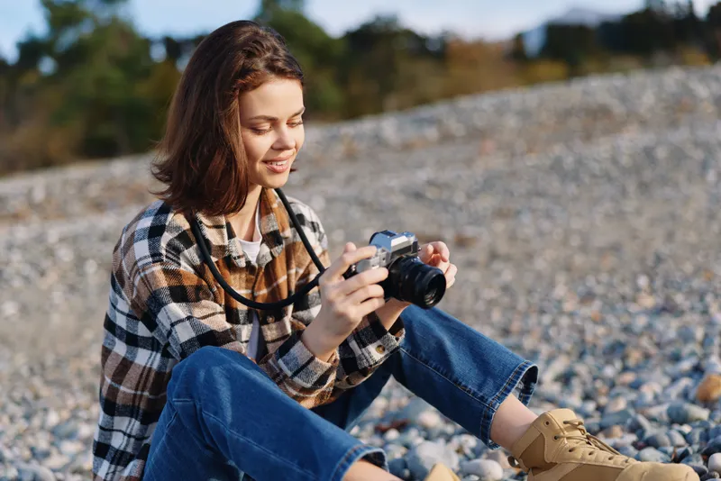 Photographer woman outdoors, sitting on pebbles with a camera, wearing a plaid shirt and jeans, capturing coastal landscape and natural light. An intimate, relaxed moment in open scenery, perfect for