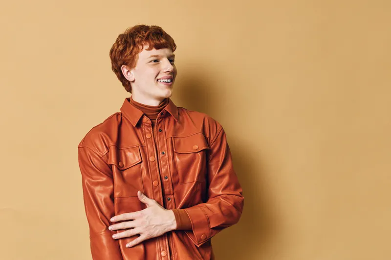 Young man with red hair wearing brown leather jacket and shirt standing smiling with hand on chest against beige background for fashion and style portrait.