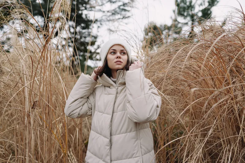 Woman in a beige puffer coat standing among tall dried grasses in a cold outdoor setting, looking upward with a calm expression softly.