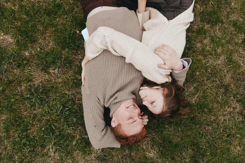 A couple lying on grass and cuddling in knit sweaters, smiling at each other. Close-up on a natural meadow setting, capturing love, warmth, and cozy lifestyle moments.
