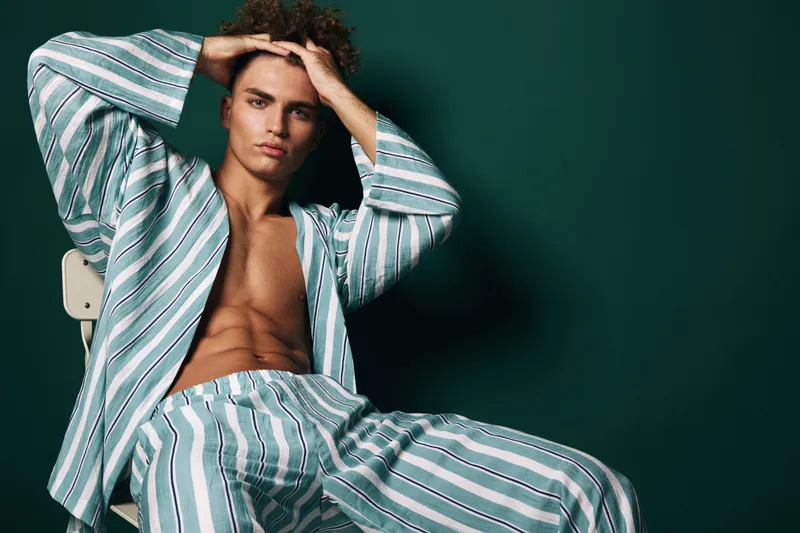 Young man with bare chest wearing striped pajamas sitting on chair with hands on head against dark background. Casual fashion, relaxed pose and confident expression in studio lighting.