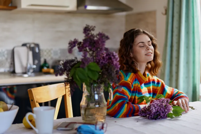 A young woman with curly brunette hair sits at a bright kitchen table arranging lilacs, her eyes closed and a peaceful smile as she enjoys a calm sunlit home moment.