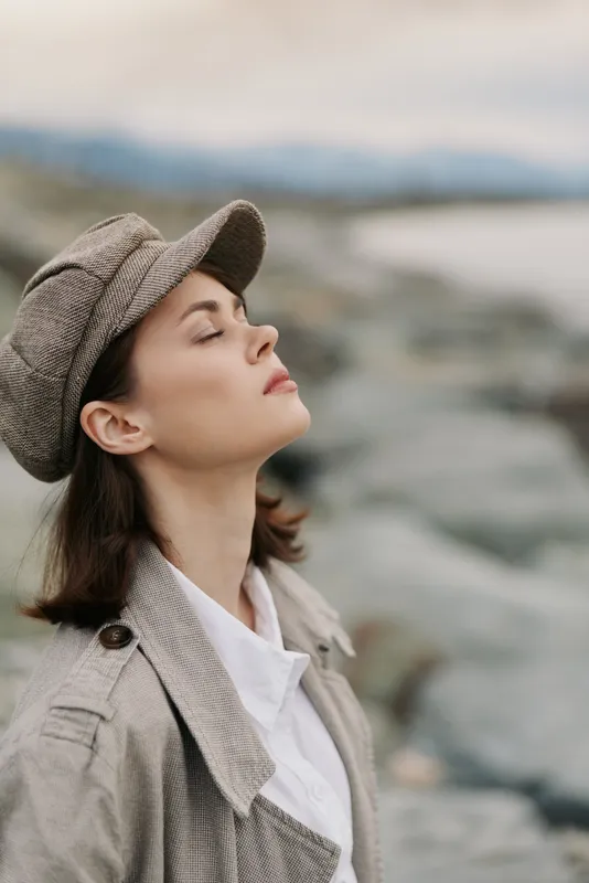 Young woman with closed eyes wearing a tweed cap and coat enjoys fresh air near rocky shore. Calm serene mood in soft natural light. Concept for relaxation, nature, and peaceful moment.