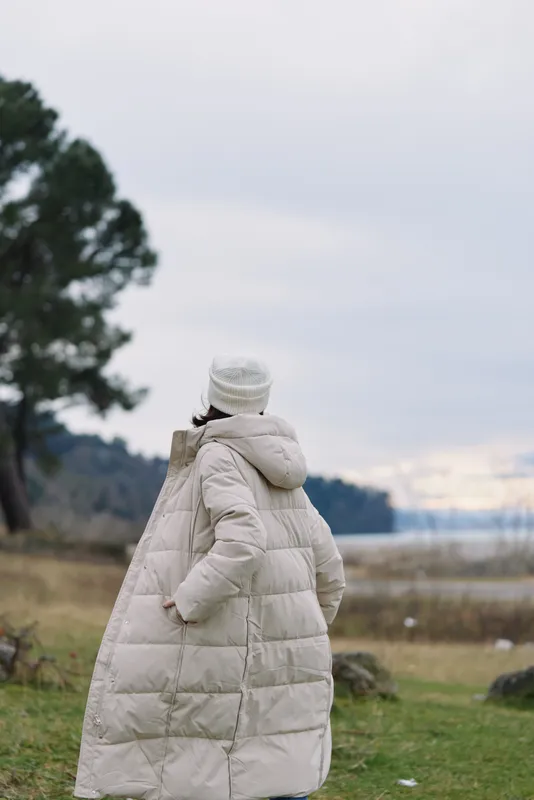 Person wearing a long beige puffer coat stands with back to camera in a chilly coastal landscape, overcast sky and distant trees frame a quiet outdoor scene.