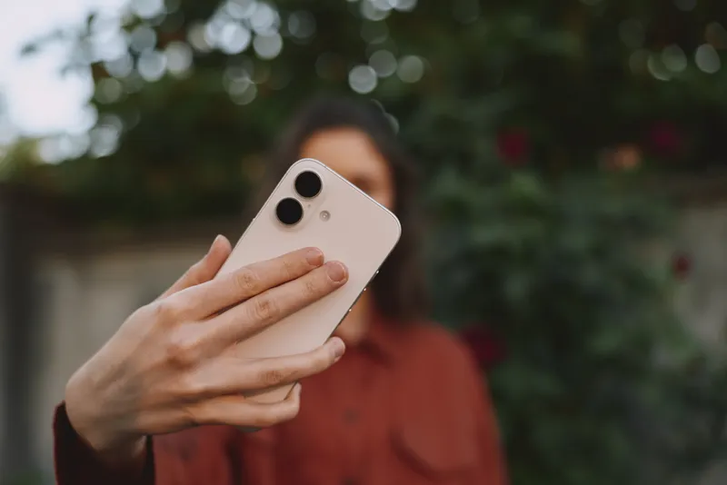 Woman lifestyle portrait taking a selfie with a smartphone outdoors, blurred garden background, casual outfit, warm natural light, candid cheerful moment, self expression