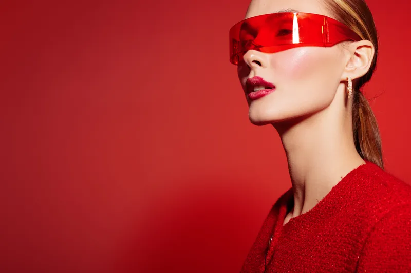 Stylish woman with red lips and futuristic red visor poses on red background. Fashion portrait with modern accessories and bold makeup. Concept of style and innovation.