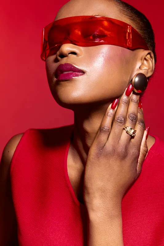 Stylish African woman with dark skin wearing red sleeveless top and large earrings poses confidently against red background. Fashion concept with red nails and glossy lips.