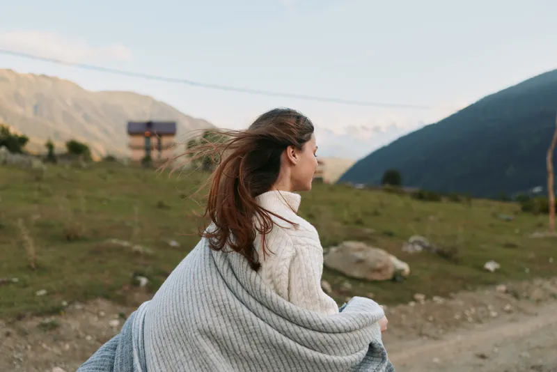 Young woman wrapped in cozy blanket enjoys fresh air outdoors looks at mountain landscape with peaceful expression on clear day in nature lifestyle concept
