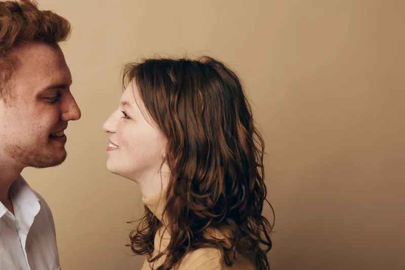 Smiling young couple with curly hair and casual clothes look at each other with affection on beige background. Concept of love, relationship, happiness, and intimacy between partners.