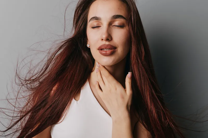 Young woman with long hair and closed eyes touching neck with hand in studio. Beauty, fashion and wellness concept with natural makeup and calm expression on face.