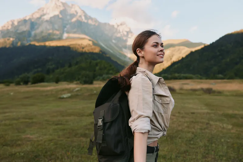 Young woman hiking outdoors with backpack, smiling and enjoying nature, mountain landscape in background, casual outfit, bright daylight, active lifestyle