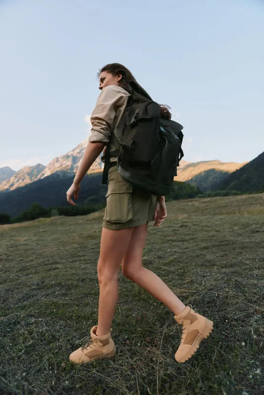 Young woman with a large backpack hikes through a mountainous landscape during golden hour, wearing shorts and boots with a focused and adventurous expression.