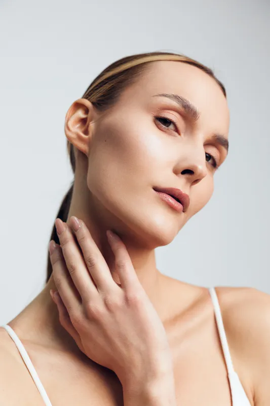 A close-up portrait of a young woman with flawless skin gently touching her neck, looking confidently at the camera. Concept for beauty, skincare, and natural elegance.