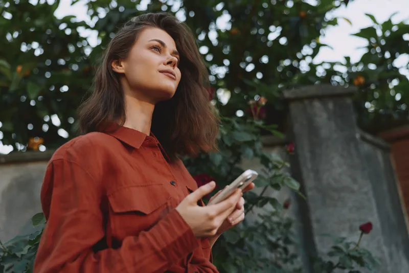 Brunette young woman in a red shirt stands outdoors, looking up while holding a smartphone, with a lush garden backdrop and a calm, thoughtful expression