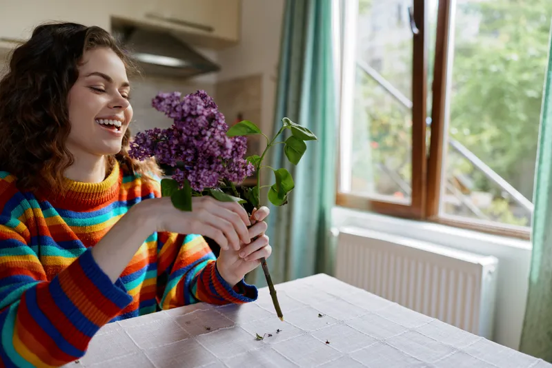 A woman enjoys a simple moment at home, arranging flowers on a table by a sunlit window. A cheerful, colorful sweater adds warmth to the cozy interior.