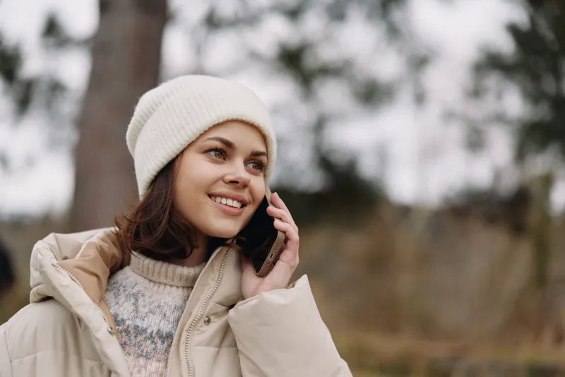 Woman outdoors wearing a beanie and warm jacket talks on her phone, smiling softly in a natural park setting during chilly weather, capturing candid everyday moments.