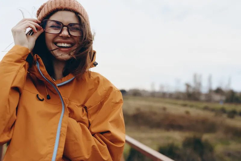 Woman lifestyle outdoors, smiling in orange jacket and knit beanie, wearing glasses, talking on a smartphone, warm autumn day, natural light, blurred field and road background, soft bokeh