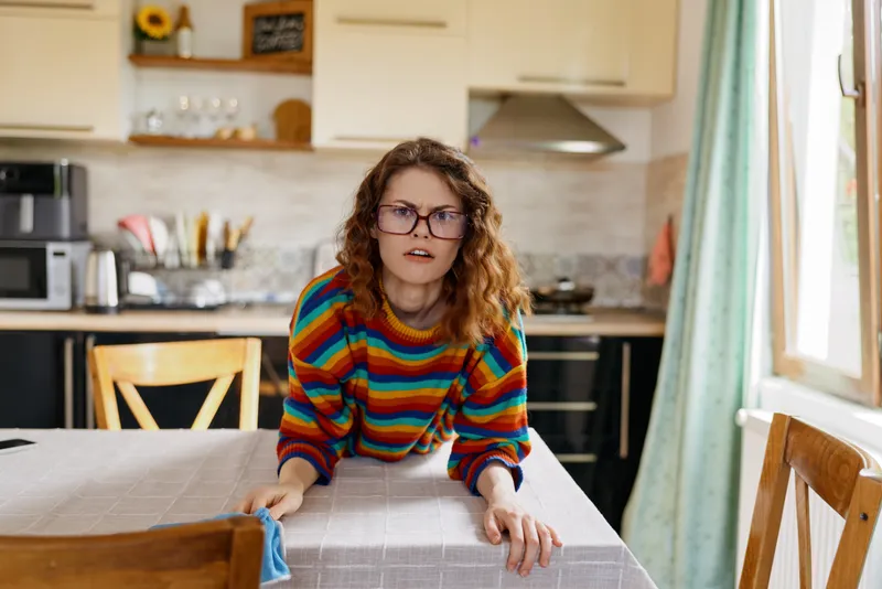 woman home kitchen lifestyle expression surprise, cheerful indoor moment with striped sweater, natural light, table foreground, casual candid vibe in a cozy kitchen