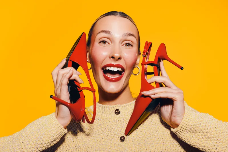 Happy young woman holds red high heel shoes near face smiling widely on bright yellow background. Concept of fashion, footwear, style, and joyful emotions in studio lighting.