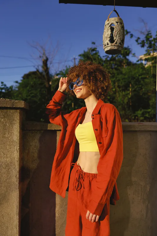 Smiling woman with curly hair wearing a yellow crop top and red-orange set outdoors. Casual lifestyle shot on a sunlit terrace, conveying confidence, fashion, and carefree summer moments. Natural