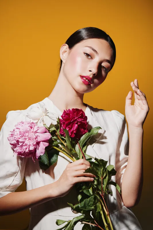 Young elegant woman with glossy black hair and red lipstick in white dress holding bright peony flowers on orange background. Creative beauty and fashion concept.