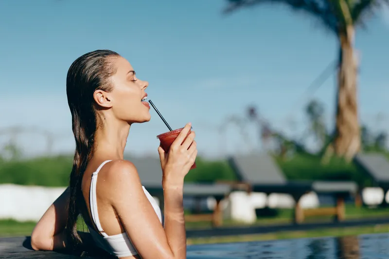 Cheerful woman relaxing by poolside, sipping a smoothie through a straw, sunlit breeze, summer vibe, tropical backdrop, casual summer style, carefree lifestyle moment