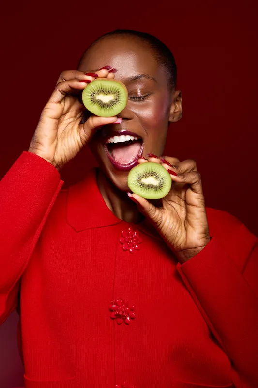 Happy African woman with dark skin in red sweater holds two kiwi halves near face smiling with open mouth on red background. Concept of healthy lifestyle, fruit, and joyful emotions.