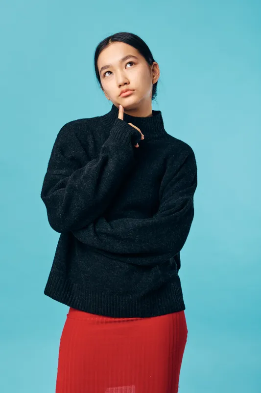 Thoughtful young woman wearing a black sweater and red skirt standing against blue background touching chin and looking up, posing with crossed arms for contemplation or decision making.