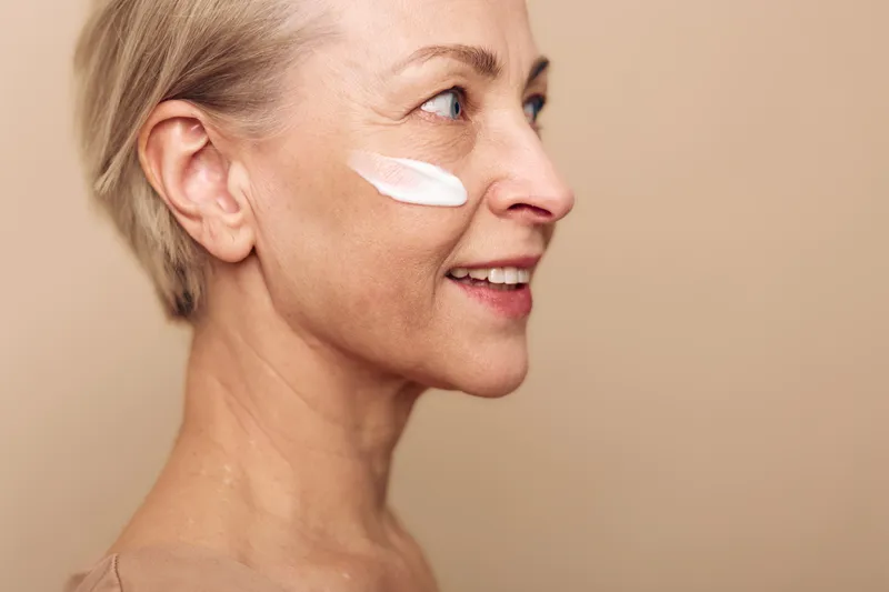 Mature woman with short hair applies face cream on cheek, smiling softly on beige background. Concept for skincare, aging skin care, beauty routine, wellness, and self-care.
