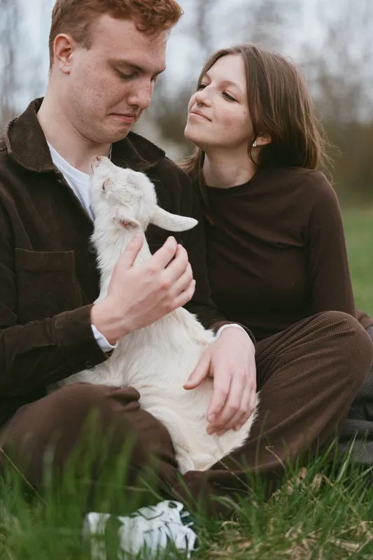Young couple sitting on grass outdoors enjoying a tender moment with a baby goat in their arms during a calm spring day in casual clothing.