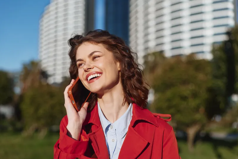 A smiling woman in a red coat talks on a mobile phone in a sunny park with an urban skyline behind. Clear concept of cheerful communication and outdoor portrait.