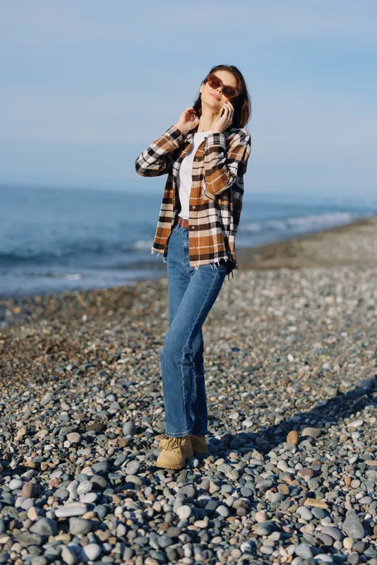 Woman on a pebble beach talking on a phone in a plaid shirt, jeans and boots, with the sea and blue sky behind in a casual outdoor lifestyle scene.