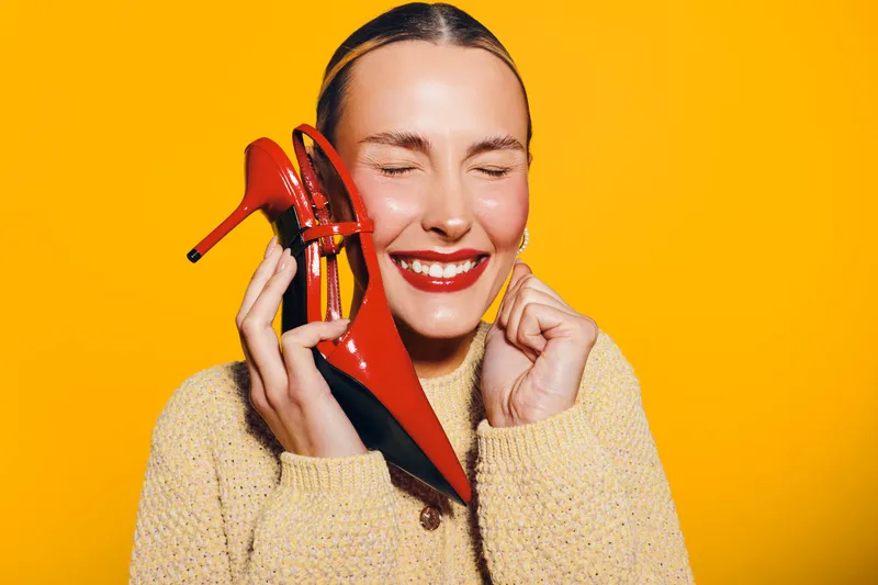 Happy woman with closed eyes holds red high heel shoe near face, smiling with bright red lipstick. Yellow background and cozy sweater create warm, joyful mood and fashion concept.