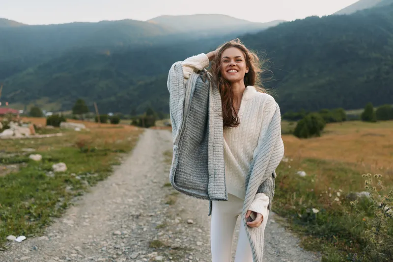 Smiling woman in cozy sweater and shawl walking on rural path with mountains in background. Outdoor lifestyle, nature, happiness, and casual fashion concept.