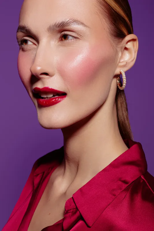 Close-up of a woman with glossy red lips and glowing skin wearing a red blouse and hoop earrings. Studio portrait on purple background with soft lighting and elegant makeup concept.