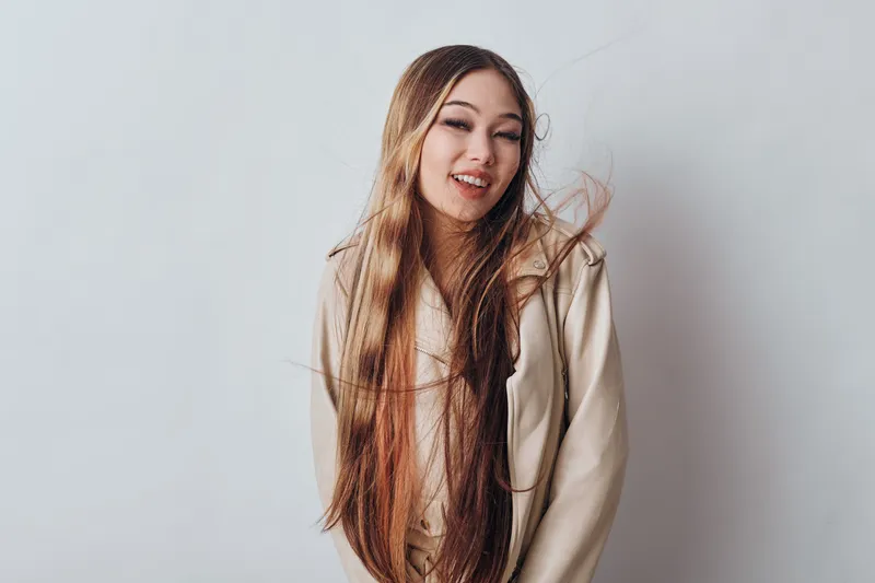 Young woman with long hair and beige jacket smiling and looking at camera on white background. Portrait of happy female with flowing hair and casual outfit in studio lighting.