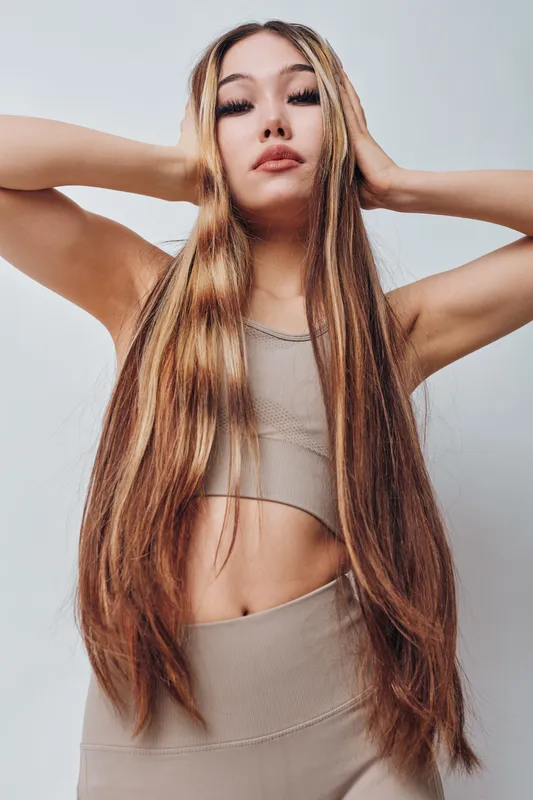 Young woman with long straight hair posing indoors with hands behind head, wearing beige top and pants. Portrait, beauty, lifestyle and fashion in studio with neutral background and confident look.