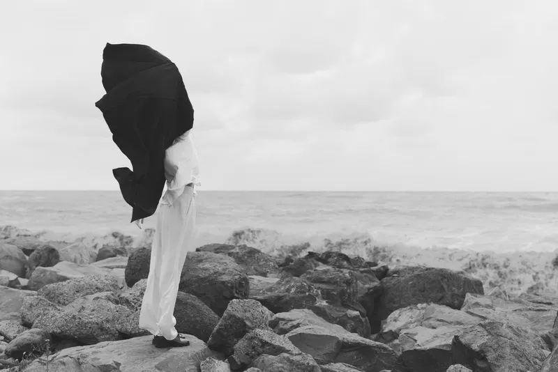 Silhouette of a figure on a rocky shore with a flowing cloak swept by wind, sea and cloudy horizon in monochrome, dramatic coastal scene and movement, capturing isolation and weathered landscape