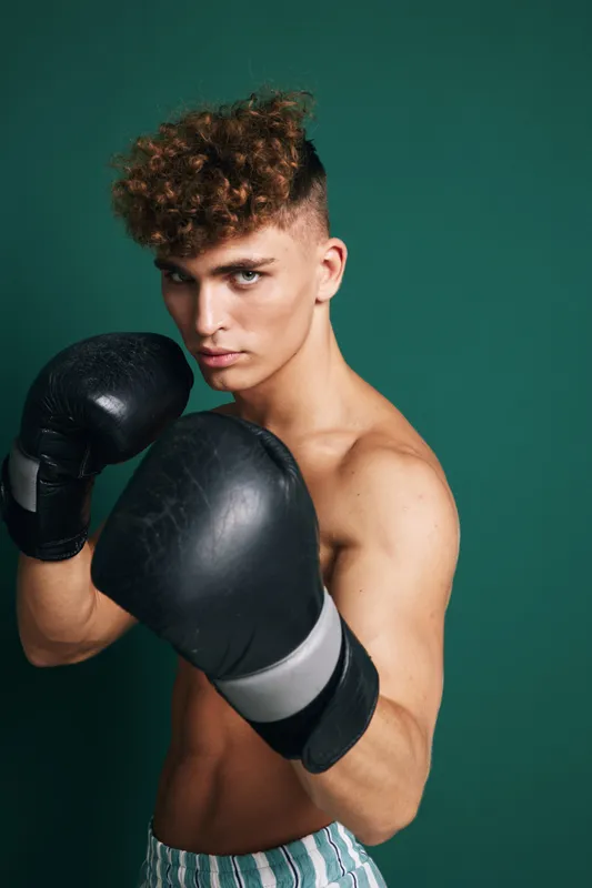 Young man with curly hair wearing boxing gloves in fighting stance, training and preparing for boxing workout against green background in studio.