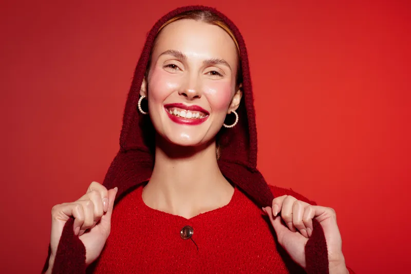 Smiling woman in red sweater and hood poses against red background. She wears hoop earrings and bright lipstick, expressing happiness and confidence in a vibrant studio setting.