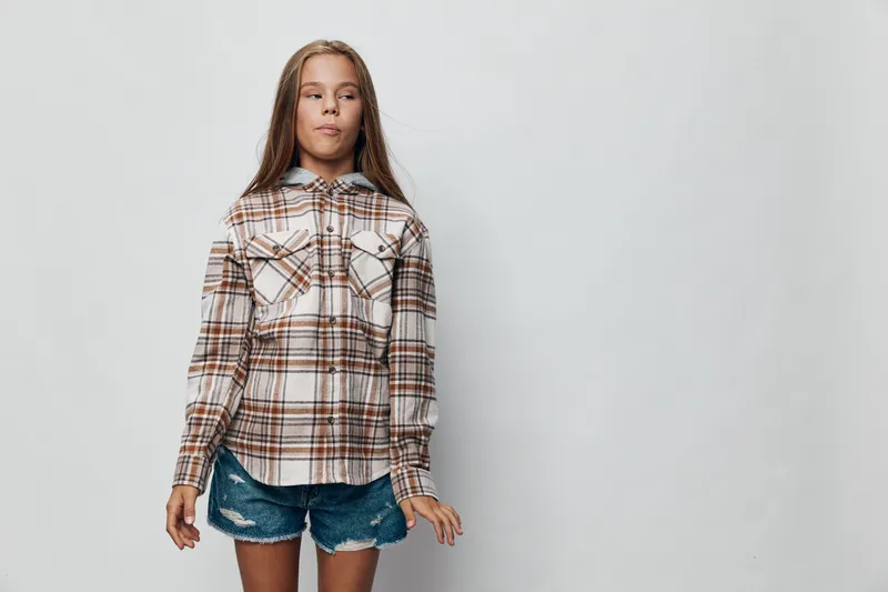 Girl standing in plaid shirt and denim shorts on light background, casual outfit and confident pose with long hair in studio for fashion and lifestyle.