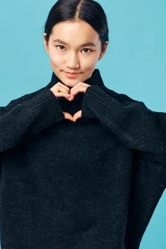 Young woman with dark hair wearing black sweater making heart shape with hands, smiling gently on blue background for love, kindness and positive emotion concept.