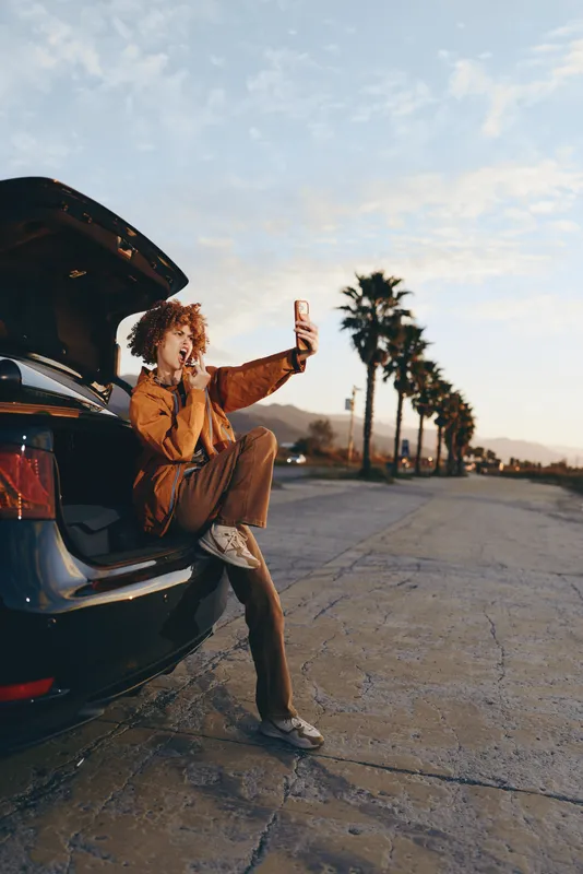 Woman lifestyle portrait outdoors beside car on quiet road at sunset, relaxed pose with curly hair blowing, casual clothing, warm light, palm trees in background, travel mood
