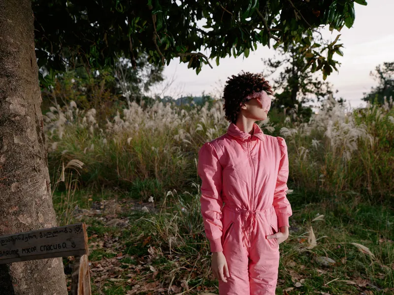 Solitary person in a pink jumpsuit poses in an outdoor portrait among a field and trees, bathed in warm sunlight for a calm, contemporary nature setting