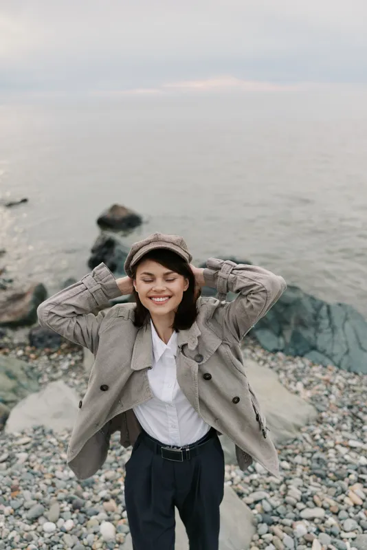 Smiling young woman in a checkered coat and hat stands on a rocky beach with closed eyes and hands behind head. Relaxed mood with ocean background on a cloudy day.