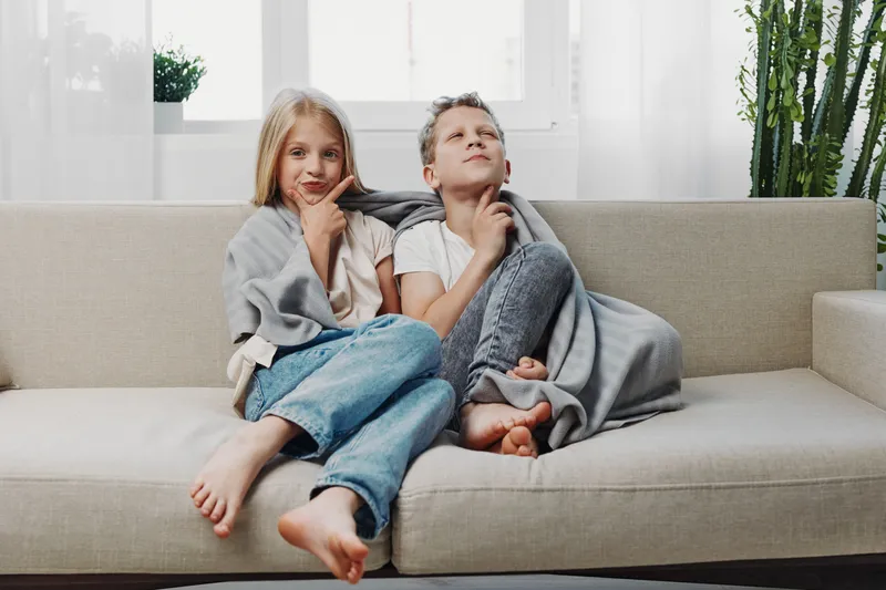 Two children sit on beige sofa with thoughtful expressions wrapped in cozy gray blanket in bright living room. Concept for family moments and childhood reflection.