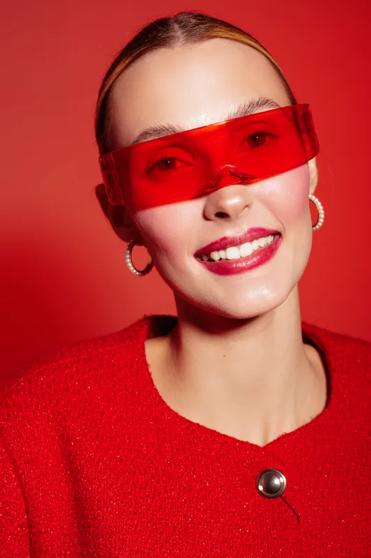 Smiling woman with red transparent glasses and red sweater poses on red background. Close-up portrait with hoop earrings and glossy lips. Concept of fashion, style, and vibrant colors.