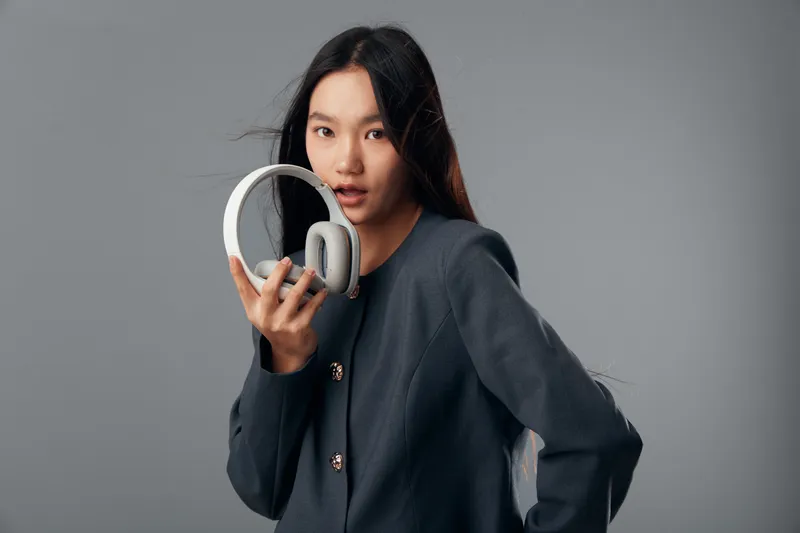 Young woman holding headphones with one hand looking at camera on grey background. Portrait of female model with long hair wearing dark jacket for music or technology concept.